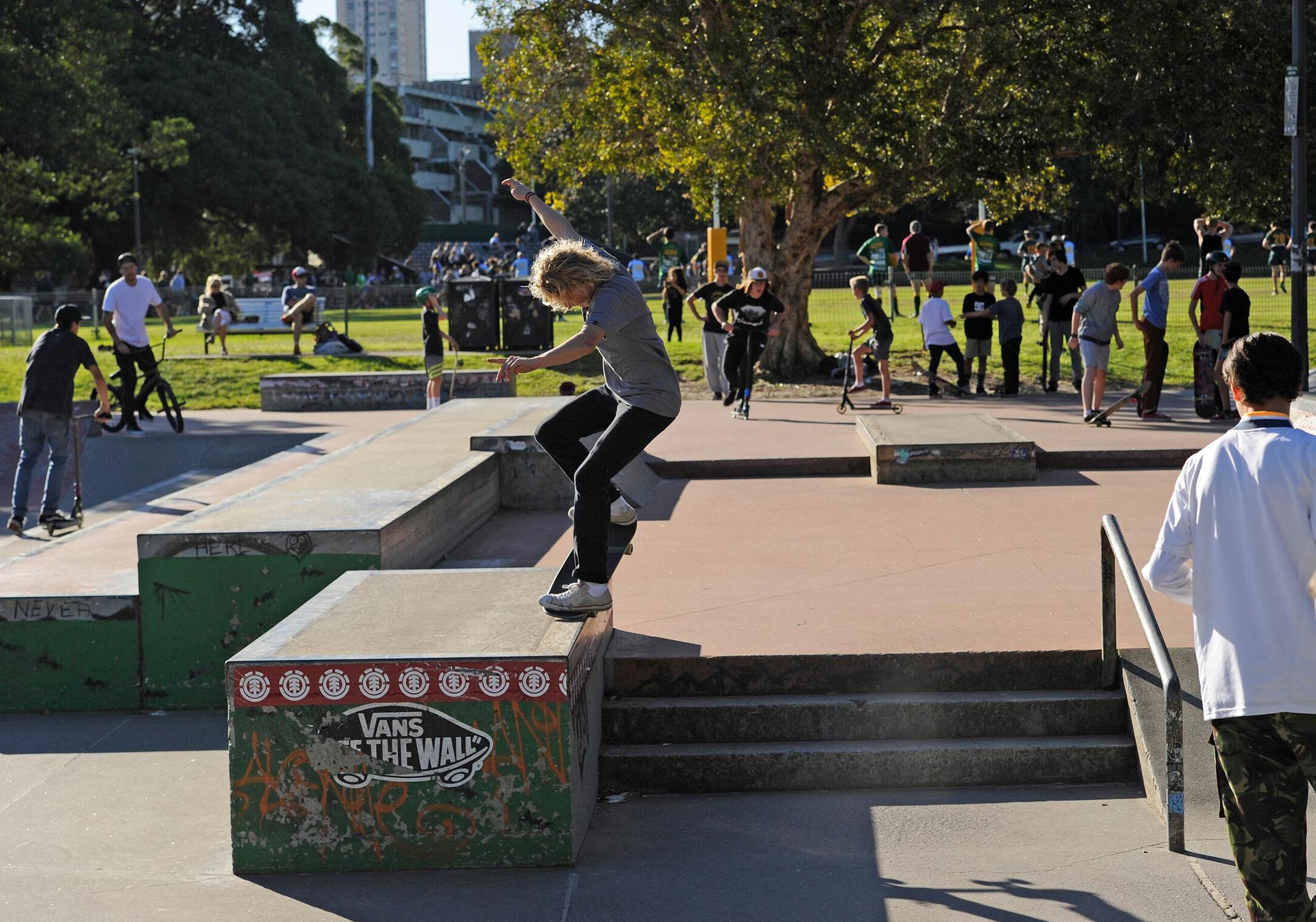 A skateboarder grinds a rail at a busy outdoor skatepark while other people ride bikes, skate, and watch in the background.